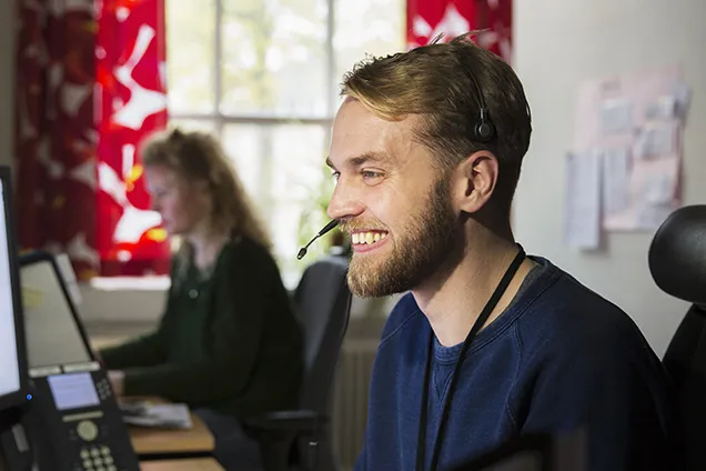 A smiling staff member in a headset in front of a computer screen.
