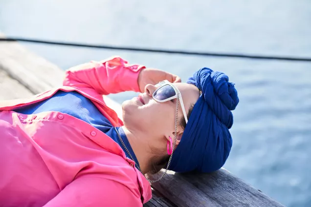 A student with sunglasses, a pink blouse and a blue turban is sunbathing on a jetty. Photographer Johan Persson.