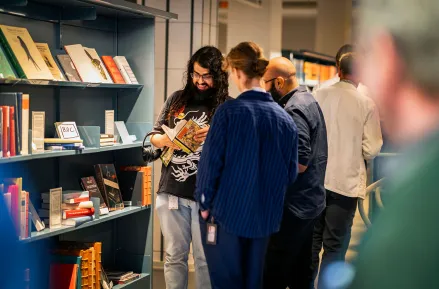 Tre personer kikar i en bok tillsammans bredvid en blå bokhylla.
